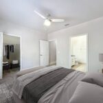 Carpeted bedroom with bed, nightstand and ceiling fan, shown with access to a walk-in closet, hallway, and en suite bathroom.