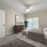 Carpeted bedroom with bed, nighstands, ceiling fan, dresser and large sliding glass door to covered patio access.